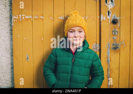 Portrait en plein air d'un drôle de petit garçon de 4-5 ans portant une veste matelassée d'hiver verte et un chapeau jaune Banque D'Images