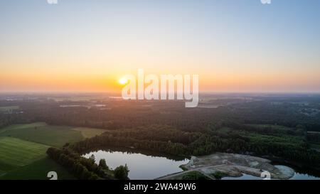 Une vue aérienne capture un lever de soleil tranquille ornant un paysage rural, projetant une lueur chaude sur de vastes champs, un lac serein, et une limite des arbres lointaine. L'horizon est orné des pastels doux de l'aube, tandis qu'une carrière ajoute une touche de contraste industriel à la scène autrement naturelle. Lever de soleil serein sur paysage rural. Photo de haute qualité Banque D'Images