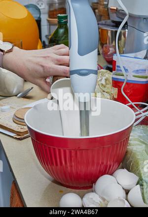 Une femme au foyer occupée est vue dans sa cuisine, fouettant habilement de la pâte à crêpes dans un bol à l'aide d'un mélangeur. L'image capture une scène domestique quotidienne o Banque D'Images