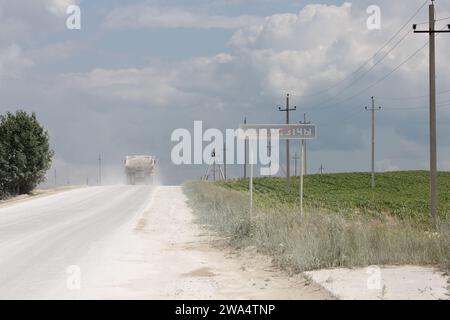 Un camion lance un nuage de poussière sur une route de campagne biélorusse, en passant un panneau de bienvenue qui fait allusion au charme rustique et à la simplicité de la région. Banque D'Images