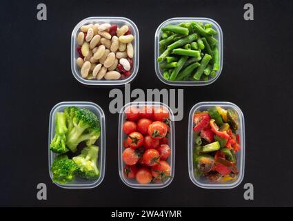 Légumes congelés dans des récipients en plastique sur fond noir. Brocoli, haricots verts, tomates cerises, haricots et poivrons congelés. Banque D'Images