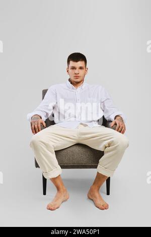 Portrait en studio complet d'un jeune homme pieds nus en chemise blanche et pantalon ivoire assis dans un fauteuil Banque D'Images