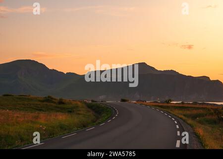 Coucher de soleil de minuit sur Lofoten avec une route courbe se dirigeant vers les montagnes, ciel clair et couleurs bleu orange foncé. Herbe verte, couleurs douces. Banque D'Images