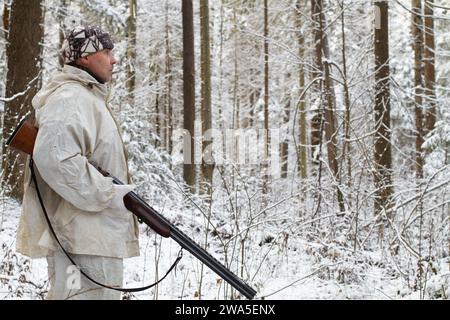 un chasseur vêtu d'un camouflage blanc avec un fusil de chasse à la main se tient dans une forêt hivernale enneigée Banque D'Images