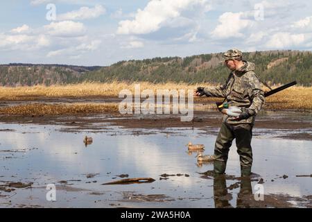 Waterfowler avec un leurre de canard dans sa main est sur l'eau peu profonde boueuse. Il se prépare pour une chasse au canard. Banque D'Images