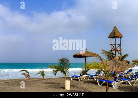 Journée plage à Boa Vista : vagues, ciel bleu avec nuages blancs, palmiers, chaises longues, parasol en rotin et tour de sauveteur en bois. Banque D'Images