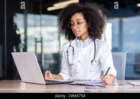 Concentré jeune femme médecin afro-américaine, étudiant praticien étudiant et travaillant en ligne sur ordinateur portable, assis au bureau dans le bureau et prenant des notes. Banque D'Images