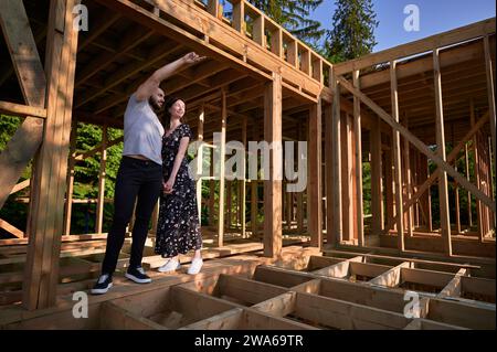 Homme et femme inspectant leur future maison de cadre en bois nichée dans les montagnes près de la forêt. Jeune couple sur le chantier en début de matinée. Concept de construction écologique contemporaine. Banque D'Images