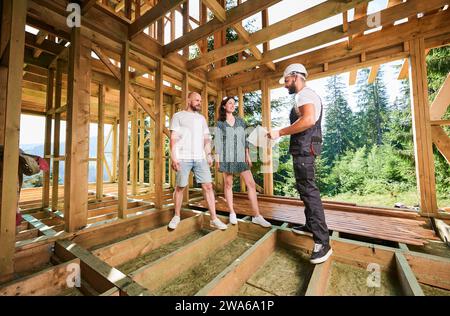 Homme travailleur avec le plan détaillé dans la main vitrines au processus de jeune couple de construction de maison à ossature en bois. Jeune couple d'investisseurs inspectant leur future demeure dans les montagnes près de la forêt. Banque D'Images