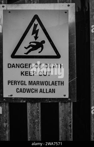 Panneau d'avertissement de danger de mort monté sur une clôture de sécurité de transformateur d'électricité. Le libellé est en anglais et en gallois. B&W. Banque D'Images