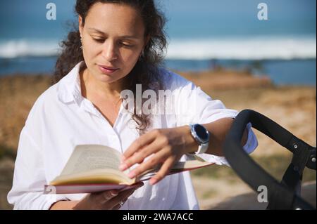 Belle frisée latino-américaine jeune femme adulte assise près de sa poussette de bébé, feuilletant à travers les pages tout en lisant roman sur la plage. D numérique Banque D'Images