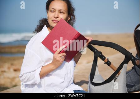 Belle femme tenant un livre dur couvert près de son visage, regardant de rêve loin, assis sur le parapet sur la plage et poussant bébé poussa, contre Un Banque D'Images