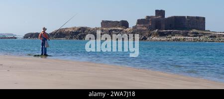 Pêcheur pêchant seul sur une plage de sable, avec des mouettes sur une petite île avec une ruine historique, dans la ville d'Essaouira, Maroc, le 2 janvier 2024 Banque D'Images