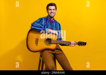 Portrait d'homme gentil joyeux positif avec poil porter noeud papillon vintage assis sur le tabouret tenir la guitare isolé sur fond de couleur jaune Banque D'Images