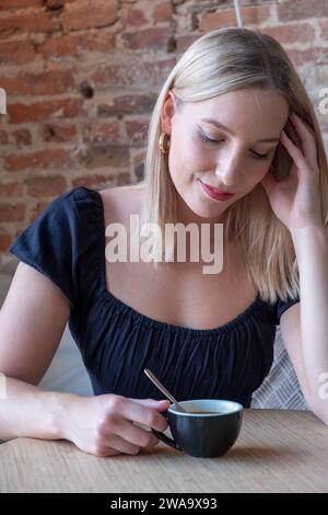 Cette image intime capture une jeune femme blonde profitant d'un moment de tranquillité avec une tasse de café. Assise à une table en bois, son regard est doucement dirigé vers le bas vers sa coupe, suggérant un moment de réflexion ou de rêverie. Le charme rustique du mur de briques en arrière-plan se juxtapose à son aspect moderne et chic. Elle porte un top noir avec manches bouffantes qui ajoute une élégance subtile à la scène. La lumière naturelle illumine doucement ses traits, soulignant ses yeux fermés et le soupçon d'un sourire, créant une atmosphère de contemplation paisible. Moment tranquille pour un Young W. Banque D'Images