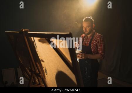 Artiste masculin talentueux utilisant énergiquement le pinceau et crée la peinture à l'huile. Studio créatif sombre avec grande toile Banque D'Images