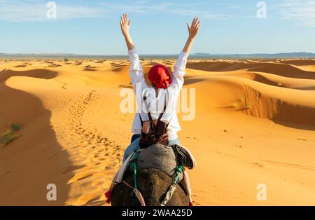 Une jeune femme les bras levés monte à dos de chameau dans les dunes du désert du Sahara. Vue de la femme de derrière, en arrière-plan de petites silhouettes Banque D'Images