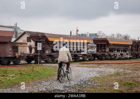 Homme plus âgé sur un vélo attendant que le train passe. Train de marchandises passant devant un passage piétonnier illégal, tandis qu'un homme avec son vélo est en attente. Banque D'Images