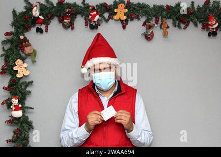 Homme latin avec masque de protection, gilet rouge et chapeau du Père Noël, avec médecine et décoration de Noël, nouveau covid-19 normal Banque D'Images