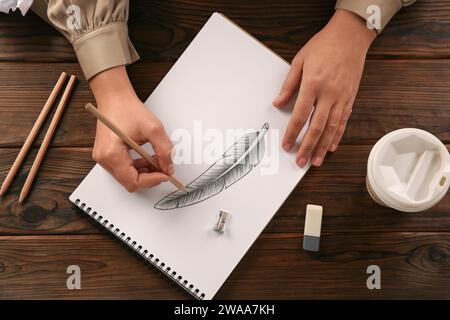 Femme dessinant plume avec crayon graphite dans le carnet de croquis à la table en bois, vue de dessus Banque D'Images