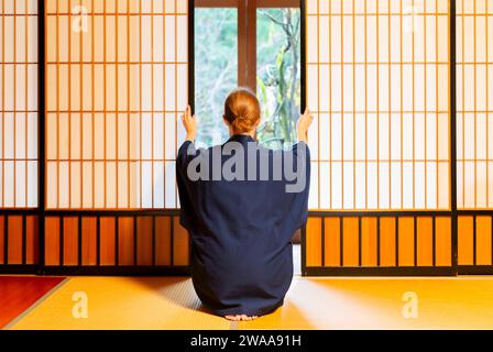 Maison traditionnelle japonaise ou ryokan avec gaijin caucasian woman en kimono et chaussettes tabi ouverture coulissante portes papier shoji assis sur tatamis cheminée Banque D'Images