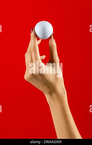 Crop anonyme femme avec manucure tenant une petite balle de golf blanche sur fond rouge. Photo concept golf. Banque D'Images