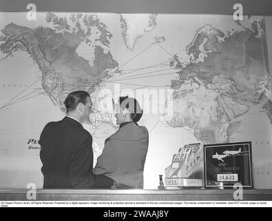 Dans les années 1950 Un jeune couple dans une agence de voyages se tient devant une carte du monde et regarde toutes les routes de vol de la compagnie aérienne américaine Pan American entre différents pays et continents. Le logo classique de Pan Am est visible sur la carte et sur le dossier du comptoir. La compagnie aérienne était la plus grande compagnie aérienne internationale aux États-Unis avec le plus de vols internationaux. 1955. Kristoffersson réf. BX51-4 Banque D'Images