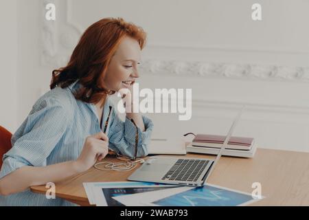 Femme aux cheveux roux embrassée dans une chemise rayée travaillant à un bureau avec ordinateur portable, ordinateur portable et tablette graphique Banque D'Images