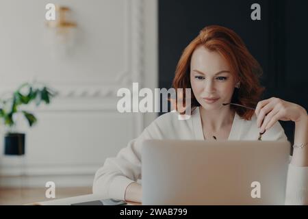 Rousse sereine avec des lunettes travaillant sur ordinateur portable, habillée d'un pull blanc chic, dans un bureau à domicile sophistiqué Banque D'Images