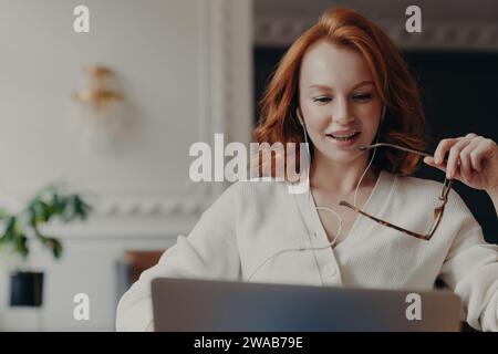 Femme aux cheveux roux en chandail blanc tenant des lunettes, souriant pendant une conversation en ligne, avec des écouteurs. Banque D'Images