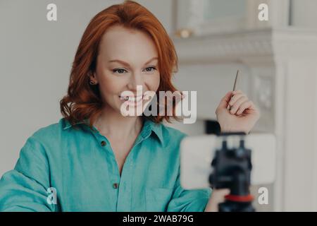Femme aux cheveux rouges vibrants enregistrant un blog avec un smartphone, stylo à la main, partageant un sourire éclatant. Banque D'Images