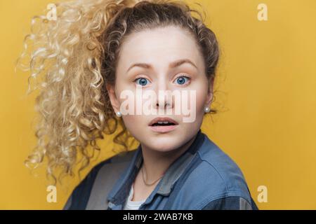 Jeune femme surprise avec des cheveux blonds bouclés en cascade, aux yeux larges dans une veste en denim, sur un fond jaune Banque D'Images