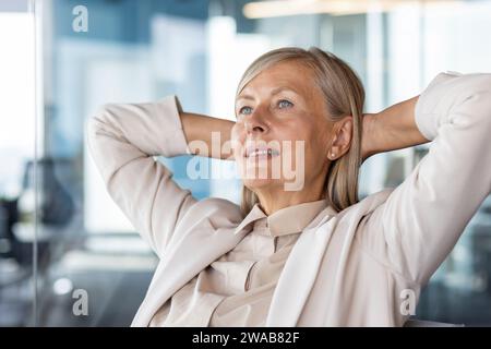 Femme d'affaires senior assise dans le bureau à la table et se reposant. Elle jeta ses mains derrière sa tête et regarda pensivement devant elle. Gros plan. Banque D'Images