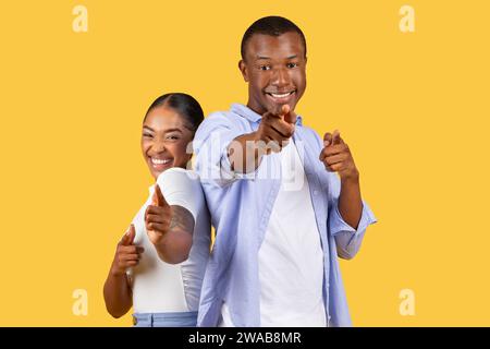 Joyeux couple noir pointant vers le spectateur, sourires engageants, jaune Banque D'Images