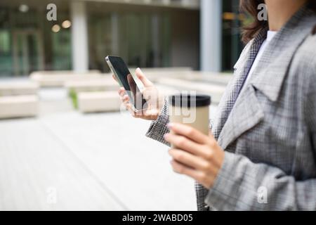 Photo recadrée d'une femme utilisant un téléphone portable tenant le café à l'extérieur Banque D'Images