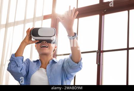 Heureuse jeune femme asiatique dans le casque VR debout par la fenêtre Banque D'Images