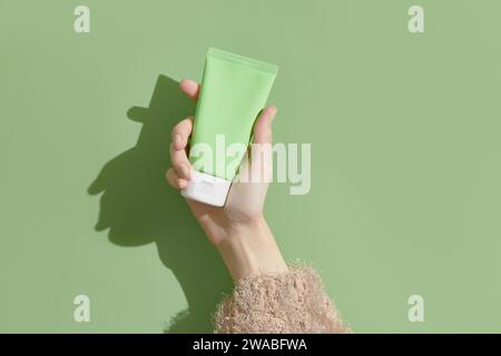 Une main de femme dans un pull tient un tube de crème sur un fond vert. Soins de beauté auto-soignés Banque D'Images