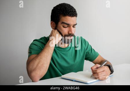 Homme sérieux avec une barbe dans un t-shirt vert écrivant dans un cahier, reposant son menton Banque D'Images