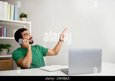 Homme barbu contemplatif dans une chemise verte, s'engageant dans une conversation téléphonique tout en faisant des gestes vers le haut Banque D'Images