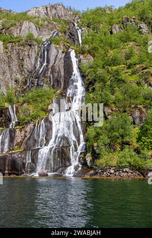 Plan vertical d'une cascade multi-ruisseaux en cascade dans le bleu profond de Trollfjorden, flanquée d'une riche végétation verte à Lofoten, Norvège Banque D'Images