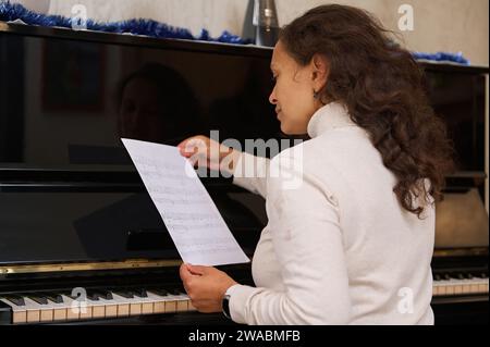 Vue arrière d'une jeune pianiste brune assise au piano et lisant des notes de musique, prête à jouer la mélodie musicale classique sur piano à queue Banque D'Images