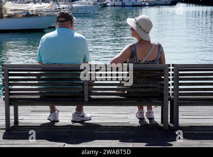 Barcelone, Espagne - 26 mai 2022 : couple senior contemplant assis sur un banc à côté de la marina, équipé de baskets, casquette et chapeau. Banque D'Images