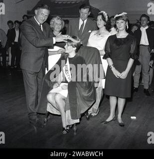 1964, historique, concours de reine de beauté.... Au milieu d'une salle de village anglaise avec des juges présents, une jeune dame, avec couronne, ceinture et cape assise sur une chaise ayant été couronnée 'Miss Prestwood'. L'ancien village de Prestwood dans les Chilterns, près de Great Missenden dans le Buckinghamshire, Angleterre, Royaume-Uni remonte à l'époque anglo-saxonne. Banque D'Images