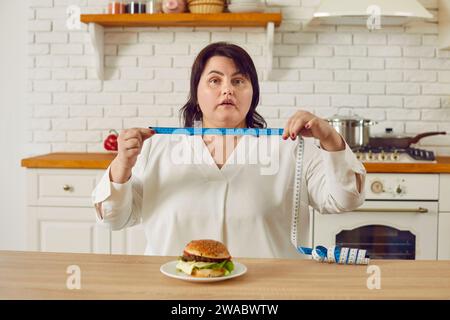 Choqué plus taille Chubby femme avec mètre ruban assis à la table devant le hamburger Banque D'Images