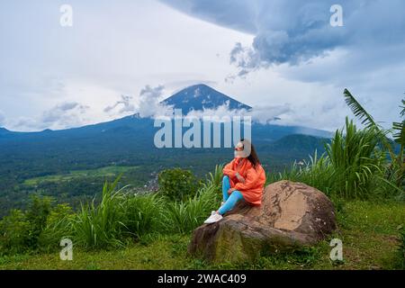 Une jeune femme est assise sur un grand rocher dans un point de vue et profite de la vue sur le volcan sacré du mont Agung caché par les nuages un jour de pluie sur l'île Banque D'Images