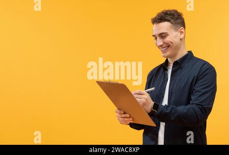 Heureux jeune homme prenant des notes dans le presse-papiers, fond jaune Banque D'Images