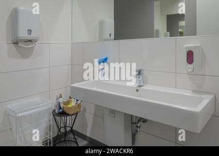 salle de bains moderne impeccable avec long lavabo blanc, sèche-mains et distributeur de savon monté sur mur carrelé. Un panier avec des produits de nettoyage se trouve à côté Banque D'Images