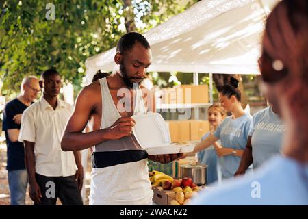 Image d'un homme afro-américain sans abri tenant sa nourriture gratuite fournie par le groupe à but non lucratif. Bénévoles travaillant pour nourrir les nécessiteux, défavorisés et défavorisés dans une banque alimentaire extérieure. Banque D'Images