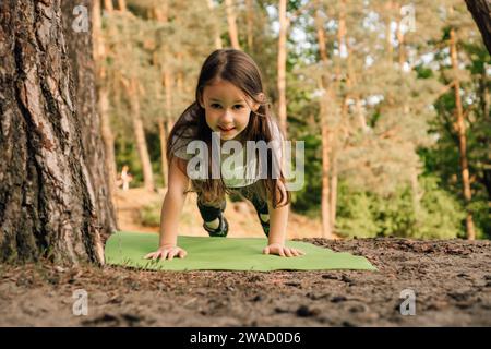 Adorable petite fille avec les cheveux fluides bruns s'exerçant et faisant de la planche sur tapis sportif vert en plein air. Athlète féminine junior s'entraînant dans la nature à proximité Banque D'Images