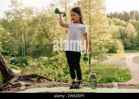 Sportif junior dynamique debout sur tapis sportif vert en plein air et s'entraîner avec de petits haltères dans les mains. Petite fille avec lifting de cheveux foncés Banque D'Images